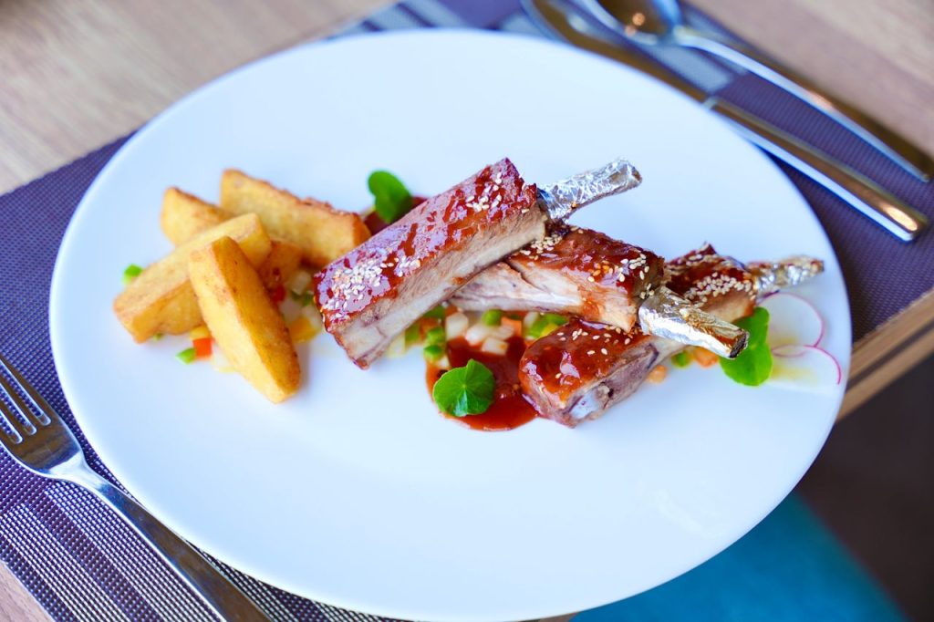 Close-up of gourmet BBQ spare ribs served with potato wedges and vegetables on a white plate.
